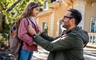 A happy dad kneels at head level with his daughter as he prepares to drop her off to school